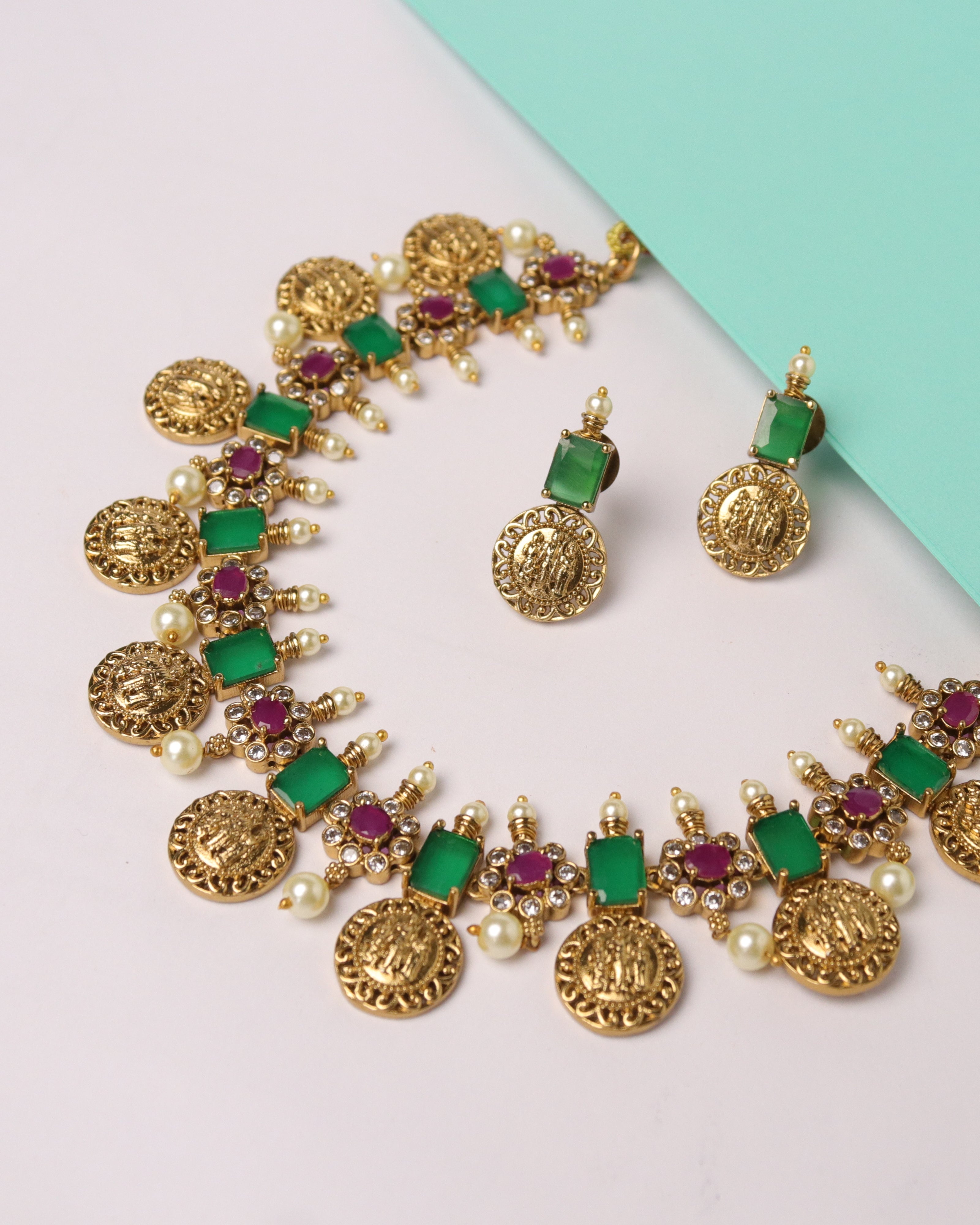 Gold jewelry set with green and red stones on a light pink and turquoise background